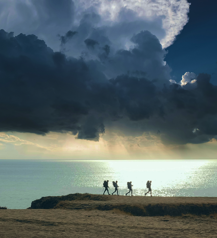 Four hikers with backpacks walking on a seaside cliff as sunlight breaks through dark storm clouds over the ocean.